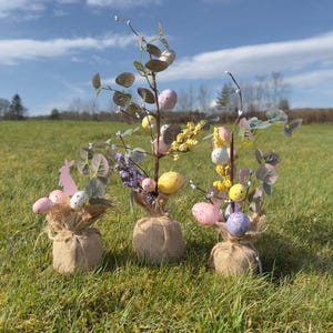 May include: Three small burlap wrapped Easter centrepieces with faux greenery, pastel coloured eggs, and purple flowers.