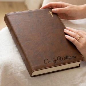 May include: A brown leather-bound book with a gold cross emblem and the name "Emily Williams" embossed on the cover. The book has cream-colored pages and is held by a person.