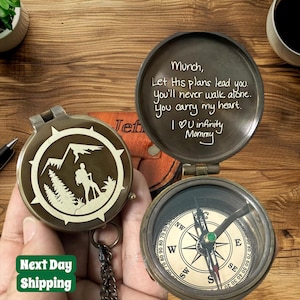 May include: A vintage-style brass compass with a mountain and hiker design. The compass is open, revealing a traditional compass face and a handwritten message inside the lid. The text "Next Day Shipping" is visible.