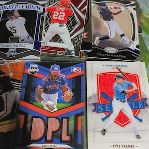 May include: A collection of baseball trading cards featuring players in action. Cards display player names, team logos, and action shots. Colors include red, blue, white, and black. Some cards have the text "HIGHER LEARNING" and "SEATTLE BASEBALL".
