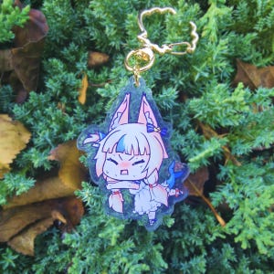 May include: A clear acrylic keychain with a cartoon character design. The character has pink and blue hair and wears a white dress. The keychain has a gold-coloured clasp, set against a backdrop of green foliage.