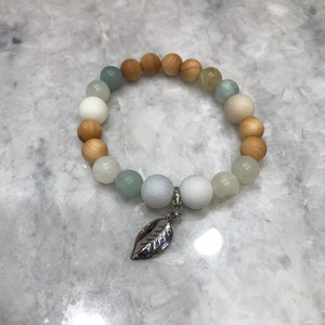 May include: A beaded bracelet featuring a mix of light-colored wooden beads, white, and pale green beads. A silver-colored leaf charm hangs from the bracelet. The bracelet is displayed on a light gray surface.