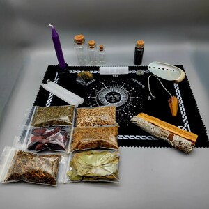 May include: A black velvet divination board with a white moon and stars design. The board includes a pendulum, a white sage smudge stick, a seashell, and six small glass bottles. The board also includes the words "Yes", "Rephrase", and "No".