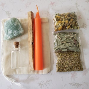 May include: A white cloth bag with a green stone, a small glass bottle with a cork top, a wooden stick, and an orange candle. There are three small plastic bags with dried herbs inside.