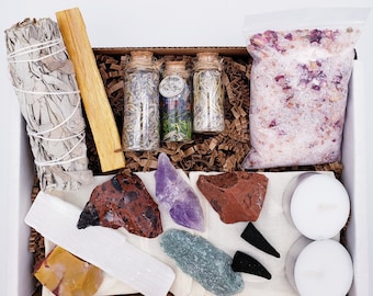May include: A gift box filled with various items for a spiritual ritual. The box contains white sage smudge sticks, palo santo wood, selenite wands, amethyst crystals, red jasper stones, green aventurine stones, rose quartz bath salts, and small glass bottles filled with herbs.