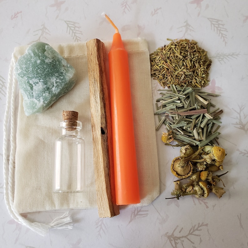 May include: A white cloth bag with a green crystal, a small glass bottle, a wooden stick, and an orange candle. The bag is surrounded by dried herbs and flowers.