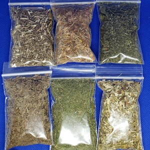 May include: Six small, clear plastic bags filled with dried herbs, arranged in two rows of three. The herbs vary in color, from light green to brown. A white candle rests at the bottom. The background is a solid blue.