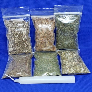 May include: Assortment of dried herbs in clear plastic bags, with a white candle on a blue background. The herbs vary in color, including green, brown, and tan. The bags are sealed with a zip closure.
