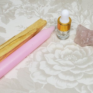 May include: A pink candle, a stick of palo santo wood, a clear glass bottle with a gold cap, and a rose quartz crystal on a floral patterned surface.