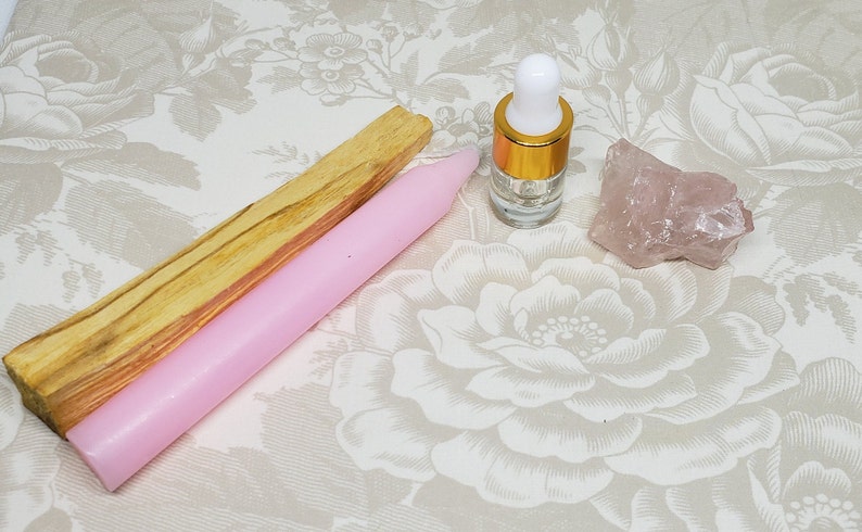 May include: A pink candle, a stick of palo santo wood, a clear glass bottle with a gold cap, and a rose quartz crystal on a floral patterned surface.