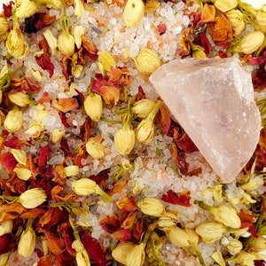 May include: A close-up of a bath salt mixture with dried flower petals and a pink crystal. The bath salt is a blend of white and pink salt crystals, and the flower petals are a mix of red, orange, and yellow.