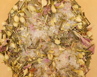 May include: A close-up of a bowl filled with bath salts, featuring a mix of dried flowers, herbs, and crystals. The salts are white, the flowers are pink, and the herbs are green.