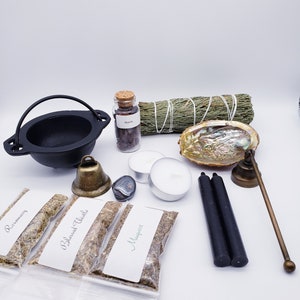 May include: A black cast iron cauldron, a small glass jar labeled "Myrrh", a bundle of sage, a seashell, two white candles, a black obsidian stone, a brass bell, two black candles, a brass incense holder, and three small bags of herbs labeled "Rosemary", "Blessed Thistle", and "Mugwort".