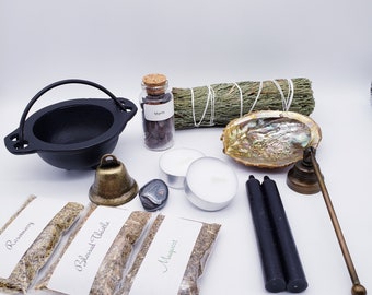 May include: A black cast iron cauldron, a small glass jar labeled "Myrrh", a bundle of sage, a seashell, two white candles, a black obsidian stone, a brass bell, two black candles, a brass incense holder, and three small bags of herbs labeled "Rosemary", "Blessed Thistle", and "Mugwort".