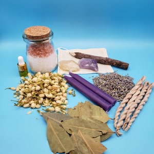 May include: A collection of items for a spiritual ritual, including a jar of pink Himalayan salt, dried lavender, dried jasmine flowers, bay leaves, a purple candle, a wooden stick, a clear crystal, a purple crystal, and a bundle of white incense.