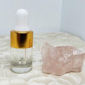May include: A clear glass bottle with a gold lid and a dropper. The bottle is filled with a clear liquid. A pink crystal is next to the bottle.