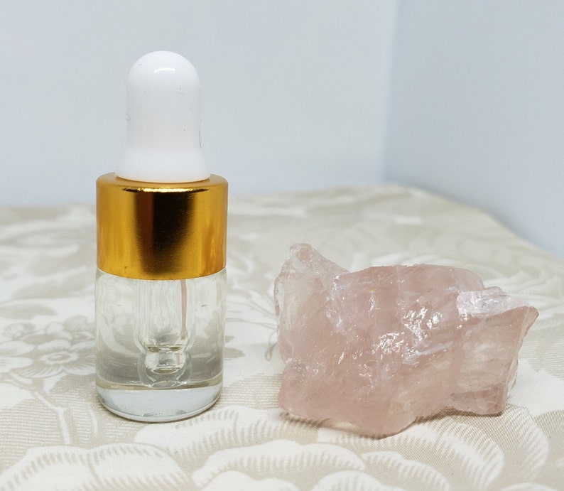 May include: A clear glass bottle with a gold lid and a dropper. The bottle is filled with a clear liquid. A pink crystal is next to the bottle.
