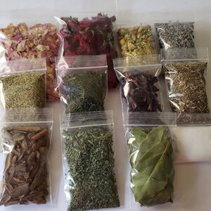 May include: Twelve clear plastic bags filled with various dried herbs and flowers. The bags are arranged in three rows of four. The herbs and flowers include rose petals, lavender, rosemary, bay leaves, and other unidentified herbs.