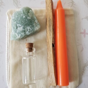 May include: A small white cloth bag containing a green crystal, a small glass bottle with a cork top, a wooden stick of incense, and an orange candle.