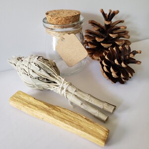 May include: A white sage smudge stick, a palo santo stick, a small glass jar with a cork lid and a brown paper tag, and two pine cones on a white surface.