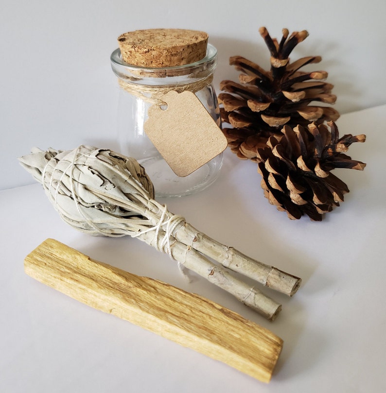 May include: A white sage smudge stick, a palo santo stick, a small glass jar with a cork lid and a brown paper tag, and two pine cones on a white surface.