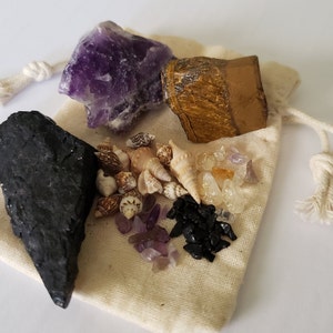 May include: A small white cloth bag filled with various crystals and seashells. The crystals include amethyst, tiger's eye, clear quartz, and black obsidian. The bag is tied with a white string.
