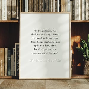 May include: A framed print with a quote from Madeline Miller's "The Song of Achilles" stands in front of a bookshelf. The quote is in black text on a white background, framed in a light wood. A small potted plant sits on the shelf.