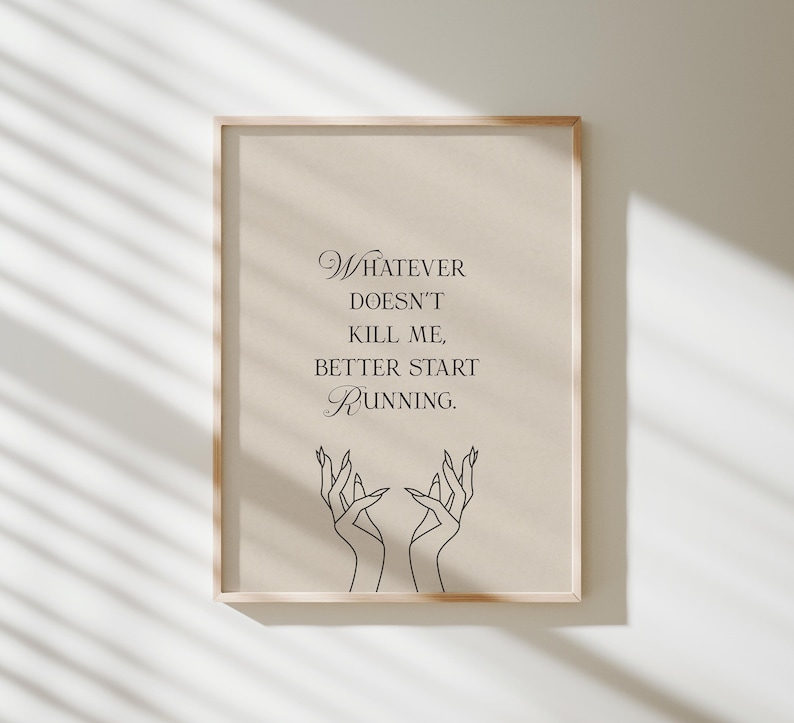 May include: Framed print with the quote "Whatever doesn't kill me, better start running." The artwork features elegant script and line art of hands reaching upwards. The frame is a light wood color, and the background is a neutral beige.