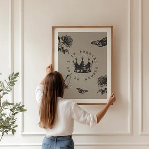 May include: A framed art print featuring a crown, roses, and butterflies in black and white. The text reads "I AM FOREVER UNDONE BY YOU". The artwork is held by a person in a white shirt and blue jeans.