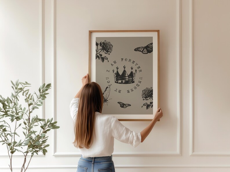 May include: A framed art print featuring a crown, roses, and butterflies in black and white. The text reads "I AM FOREVER UNDONE BY YOU". The artwork is held by a person in a white shirt and blue jeans.