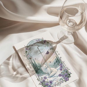 Puede incluir: Una tarjeta de felicitaci&oacute;n con un zorro blanco descansando en un paisaje nevado con una espada y el texto "she is moonlight". La tarjeta est&aacute; rodeada de flores moradas y mariposas. Una copa de vino est&aacute; en el fondo.