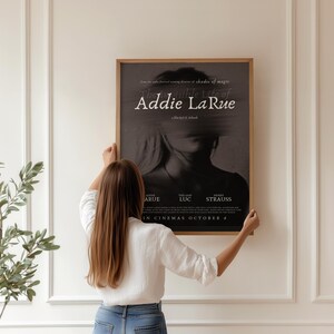 K&ouml;nnte beinhalten: Eine Frau h&auml;ngt ein Filmplakat an einer wei&szlig;en Wand. Das Plakat ist f&uuml;r einen Film namens "Addie La Rue" und zeigt ein Schwarzwei&szlig;bild eines Frauen Gesichts. Das Plakat enth&auml;lt auch die Namen der Besetzung und der Crew sowie das Erscheinungsdatum.