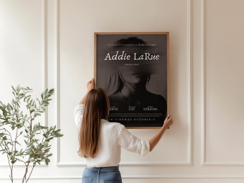 K&ouml;nnte beinhalten: Eine Frau h&auml;ngt ein Filmplakat an einer wei&szlig;en Wand. Das Plakat ist f&uuml;r einen Film namens "Addie La Rue" und zeigt ein Schwarzwei&szlig;bild eines Frauen Gesichts. Das Plakat enth&auml;lt auch die Namen der Besetzung und der Crew sowie das Erscheinungsdatum.
