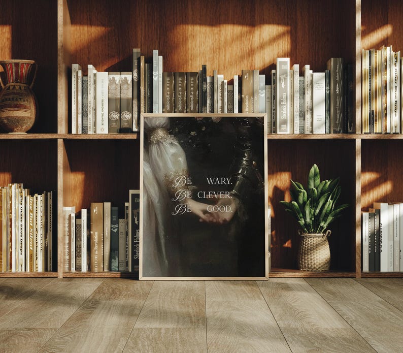 Puede incluir: Una impresi&oacute;n enmarcada con el texto "Be wary. Be clever. Be good." sobre un fondo oscuro. La impresi&oacute;n se muestra en un suelo de madera frente a una estanter&iacute;a llena de libros y una peque&ntilde;a planta en maceta.