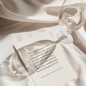 May include: A clear wine glass and a book with text on a white surface. The book's text reads, "Does such a thing as 'the fatal flaw,' that showy dark crack running down the middle of a life..." The book is titled "THE SECRET HISTORY."
