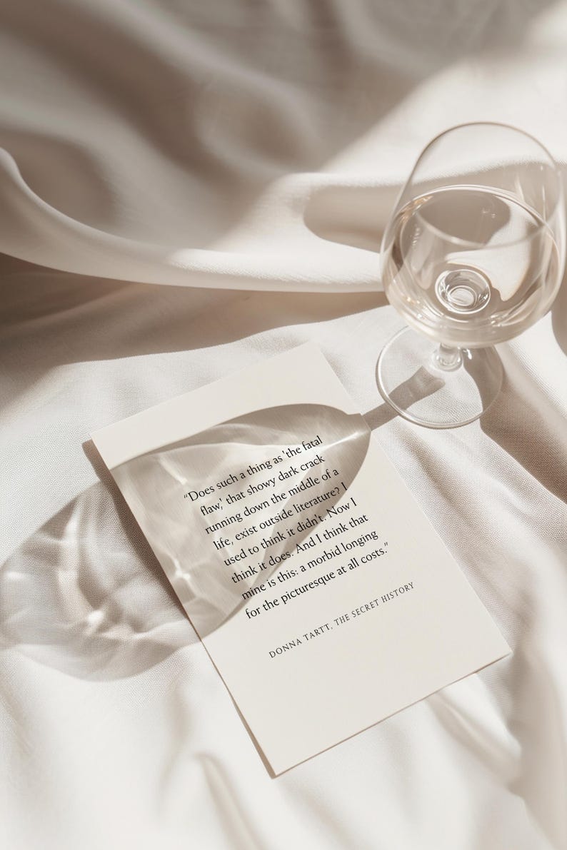 May include: A clear wine glass and a book with text on a white surface. The book's text reads, "Does such a thing as 'the fatal flaw,' that showy dark crack running down the middle of a life..." The book is titled "THE SECRET HISTORY."