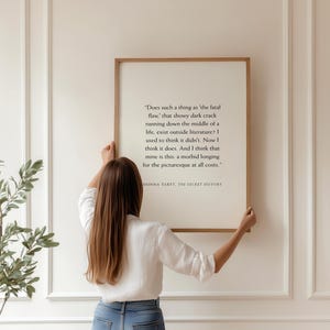 May include: A framed print with a quote from Donna Tartt's "The Secret History." The text is in black on a cream background, framed in a light brown wood. A person is holding the framed print.