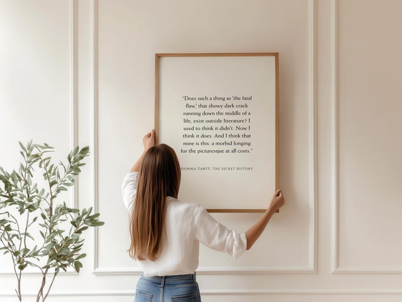 May include: A framed print with a quote from Donna Tartt's "The Secret History." The text is in black on a cream background, framed in a light brown wood. A person is holding the framed print.