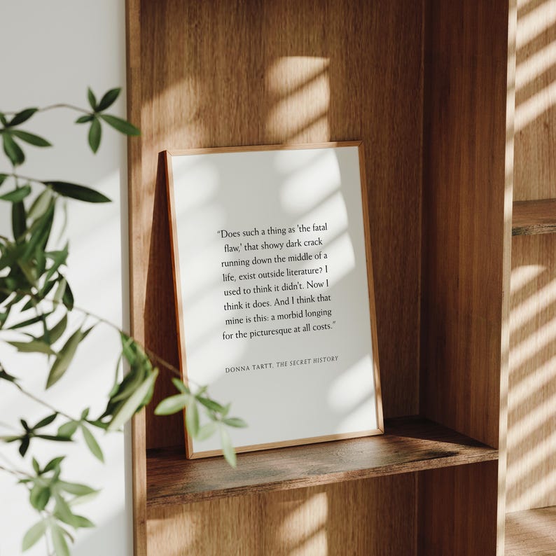 May include: A framed print with a quote from Donna Tartt's 'The Secret History' on a wooden shelf. The text is in black on a white background, framed in a thin, light-colored wood. The shelf is made of dark wood.
