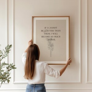 May include: A framed print with the quote "IF I CANNOT BE BETTER THAN THEM, I WILL BECOME SO MUCH WORSE" in black text. The print has a beige background and a floral illustration. The frame is a light brown colour.