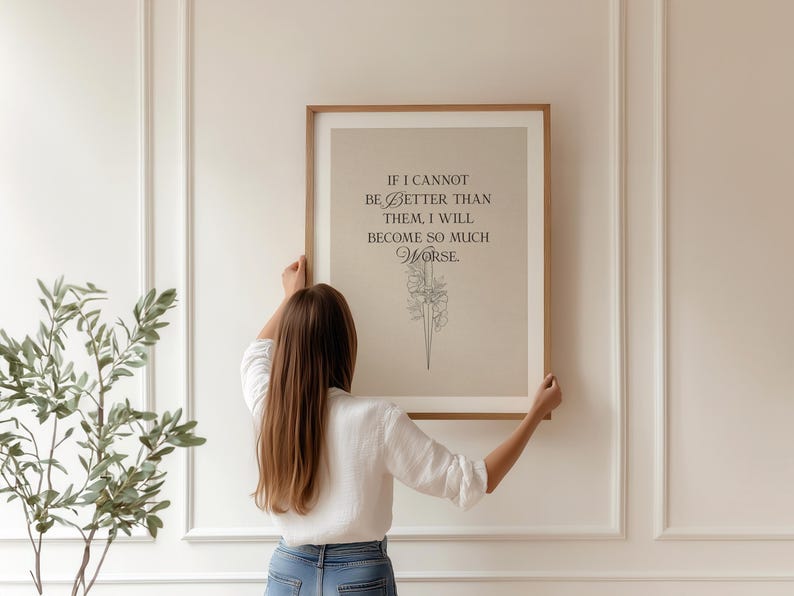 May include: A framed print with the quote "IF I CANNOT BE BETTER THAN THEM, I WILL BECOME SO MUCH WORSE" in black text. The print has a beige background and a floral illustration. The frame is a light brown colour.