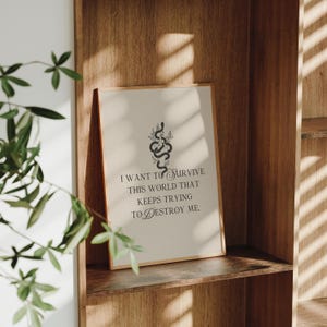 May include: A framed print with a beige background and a gold frame. The print features a black snake design with botanical elements and the text: "I WANT TO SURVIVE THIS WORLD THAT KEEPS TRYING TO DESTROY ME."