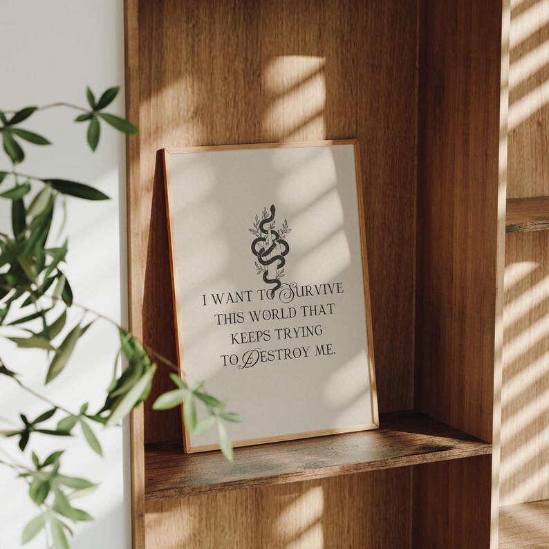 May include: A framed print with a beige background and a gold frame. The print features a black snake design with botanical elements and the text: "I WANT TO SURVIVE THIS WORLD THAT KEEPS TRYING TO DESTROY ME."