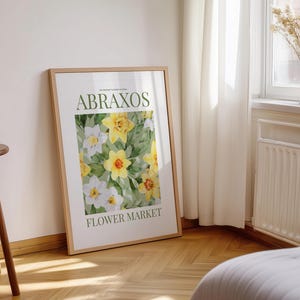 Puede incluir: Un cuadro enmarcado con un dise&ntilde;o floral de flores amarillas y blancas. El texto "ABRAXOS FLOWER MARKET" es visible. El marco es de color marr&oacute;n claro. El cuadro est&aacute; apoyado contra una pared blanca.