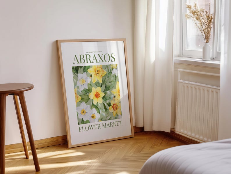 Puede incluir: Un cuadro enmarcado con un dise&ntilde;o floral de flores amarillas y blancas. El texto "ABRAXOS FLOWER MARKET" es visible. El marco es de color marr&oacute;n claro. El cuadro est&aacute; apoyado contra una pared blanca.