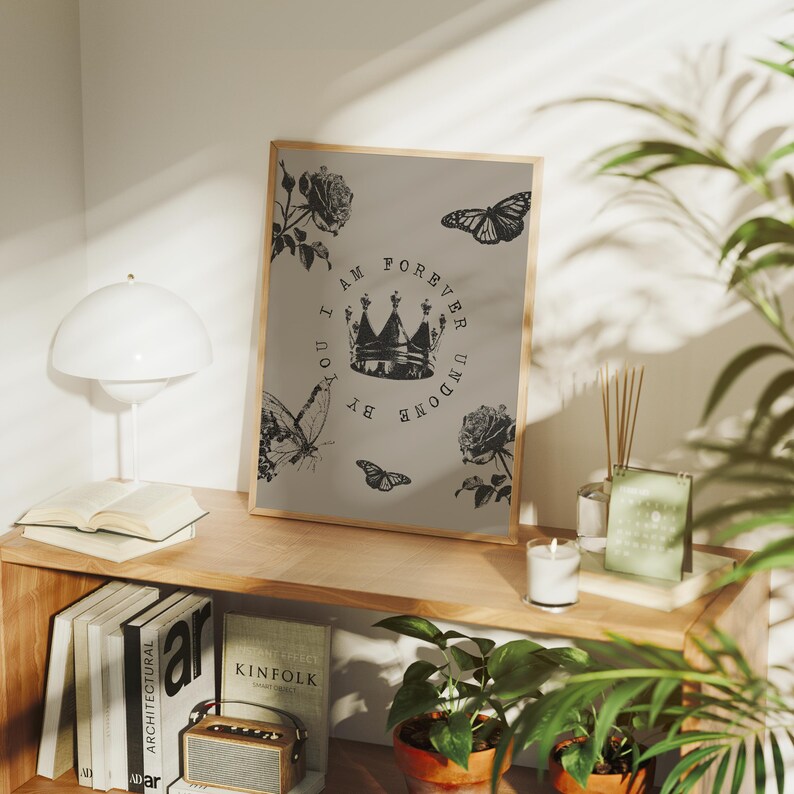 May include: A framed print with a light brown background featuring a crown, butterflies, and roses. The text reads "I AM FOREVER UNDONE BY YOU." The artwork is displayed on a wooden shelf with books, a lamp, and plants.