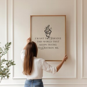 May include: A framed art print with a light background and a wooden frame. The print features a black snake design with the quote, "I want to survive this world that keeps trying to destroy me." A person is holding the framed art.