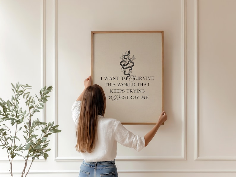 May include: A framed art print with a light background and a wooden frame. The print features a black snake design with the quote, "I want to survive this world that keeps trying to destroy me." A person is holding the framed art.