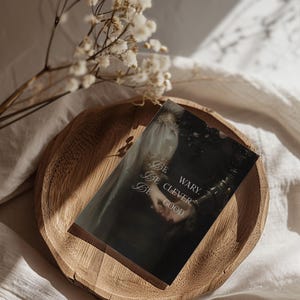 Puede incluir: Una postal oscura de estilo vintage con el texto "Be wary. Be clever. Be good." se muestra en un plato de madera. La postal presenta una imagen oscura y rom&aacute;ntica de una persona con un vestido blanco. Flores secas en el fondo.