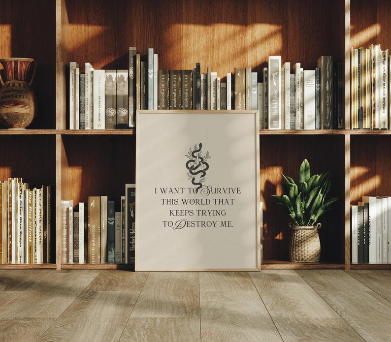 May include: A framed print with the text "I want to survive this world that keeps trying to destroy me." The print features a black snake and floral design. The print is displayed on a wooden floor in front of a bookshelf.